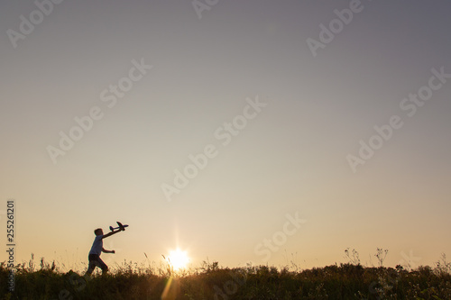 Black silhouette of cute happy cheerful child running fastly along grassy hill at countryside holding big toy plane in hand. Boy playing during sunset time in evening. Horizontal color photography.