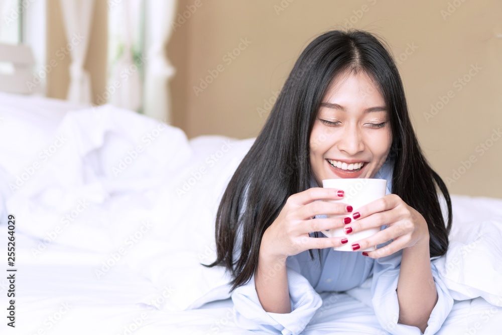 Happy smiling Asian young woman lying holding a cup of coffee on white bed in the morning at home.
