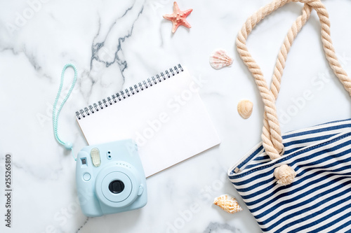 Summer travel composition on a marble background. Women's desk with instant photo camera, striped beach bag, seashells and blank notepad. Lifestyle of traveler, beauty blogger, digital nomad, hipster.