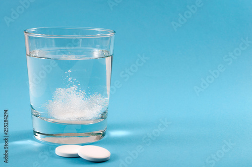 Recovering from a hangover and nursing a headache with aspirin concept with effervescent drink tablet dissolving in water with two tablets outside the glass isolated on blue background with copy space photo