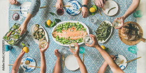 Mediterranean style dinner. Flat-lay of table with cooked salmon, starters, bread over blue table cloth with hands holding drinks, sharing food, top view, wide composition. Holiday party concept