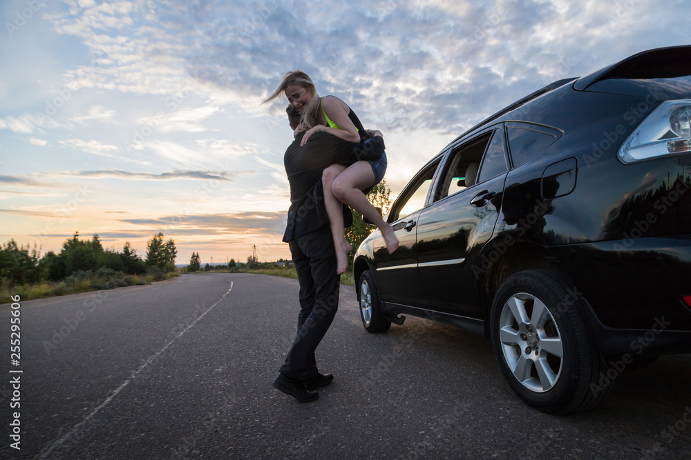 Guy carrying the girl on the road near the car in a summer evening
