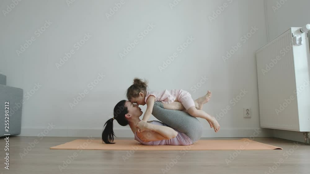 YOGA: Mother lies on yoga mat with little daughter on her legs at home - Side view YOGA: Mother lies on yoga mat with little daughter on her legs at home - Side view
