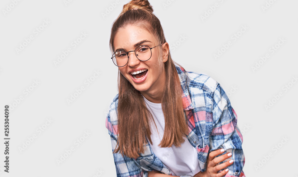 Happy beautiful positive young woman dressed in casual outfit, wearing round transparent eyewear with pleasant broadly smile, looking to the camera and posing over white studio wall. People emotions