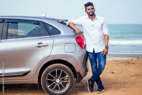 Indian businessman standing near car outdoors on sea beach summer good day.a man in a white shirt and snow-white smile rejoicing buying a new car enjoying a vacation by the ocean © yurakrasil