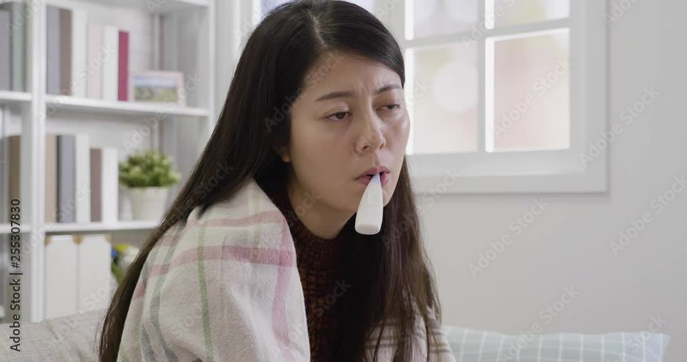 ill woman with thermometer in her mouth sitting on sofa feeling tired uncomfortable. unhealthy asian woman cold cover body with blanket. Sick woman with seasonal flu measuring body temperature. ill woman with thermometer in her mouth sitting on sofa feeling tired uncomfortable. unhealthy asian woman cold cover body with blanket. Sick woman with seasonal flu measuring body temperature.