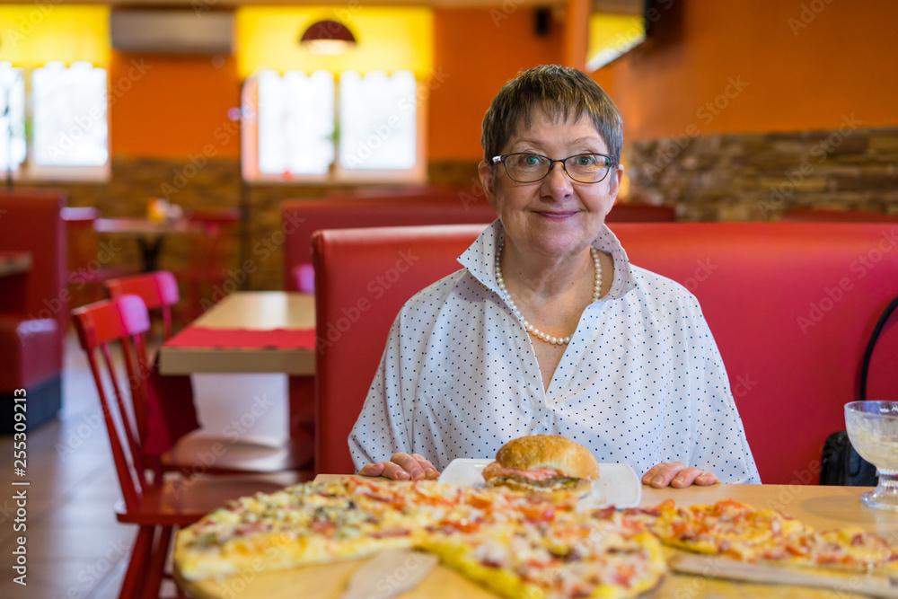 The grandmother in the cafe-restaurant ordered a bunch of food. Pizza, sushi, rolls, hamburger ... An elderly woman is happy with so much fast food.