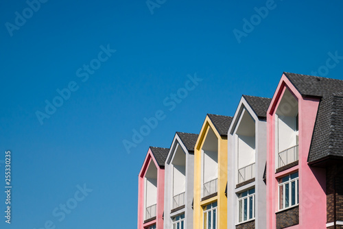 Fototapeta Rows of triangular elements of roofline covered with shingle, colorful pink and yellow house against blue sky