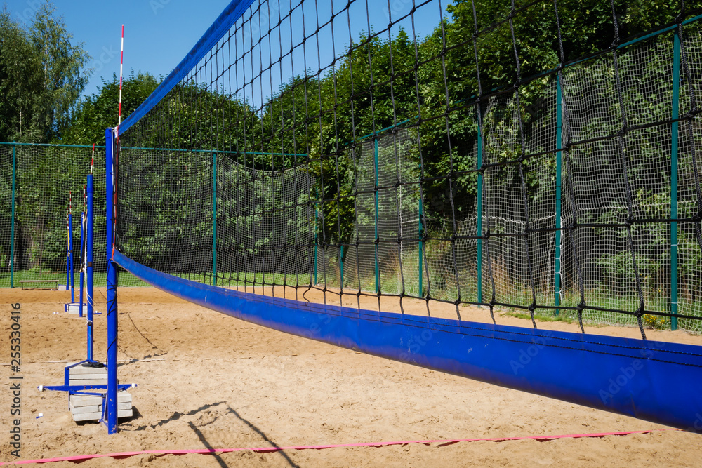 Close up of volleyball nets on blue poles on empty sand court with lush green trees in background on summer day Close up of volleyball nets on blue poles on empty sand court with lush green trees in background on summer day