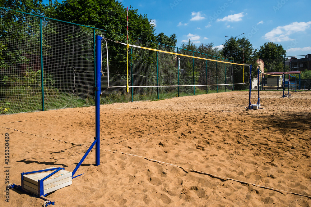 Volleyball nets on blue poles with concrete weights on empty sand court surrounded with lush green trees on summer day