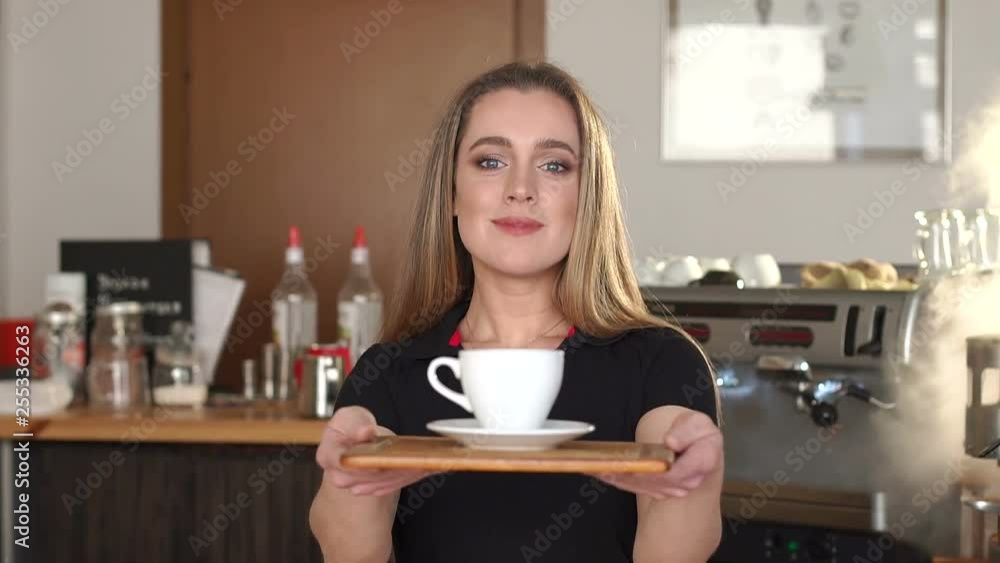 Beautiful female barista is holding a cup, looking at camera and smiling while standing near the bar counter in cafe. Portrait of a cute young Barista girl, she smiles and offers you coffee. Beautiful female barista is holding a cup, looking at camera and smiling while standing near the bar counter in cafe. Portrait of a cute young Barista girl, she smiles and offers you coffee.