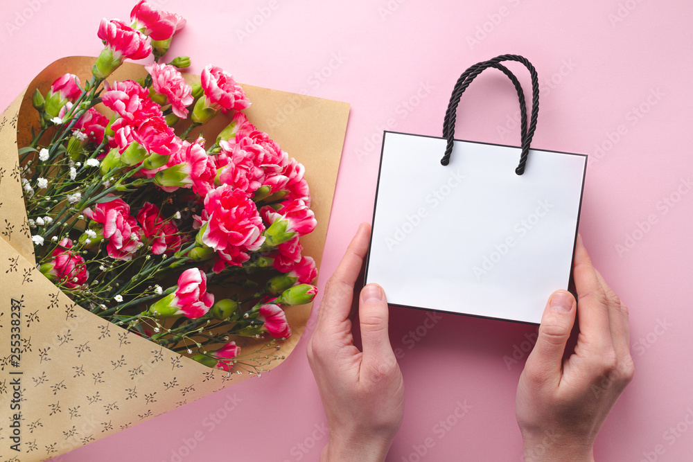 Obraz premium Bouquet of fresh carnations and a paper white gift bag on pink background. Receiving gifts and flowers from loved ones