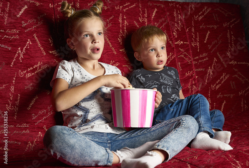 Children's cinema: A girl and a boy watch a movie at home on a big red sofa in the dark and eat popcorn.