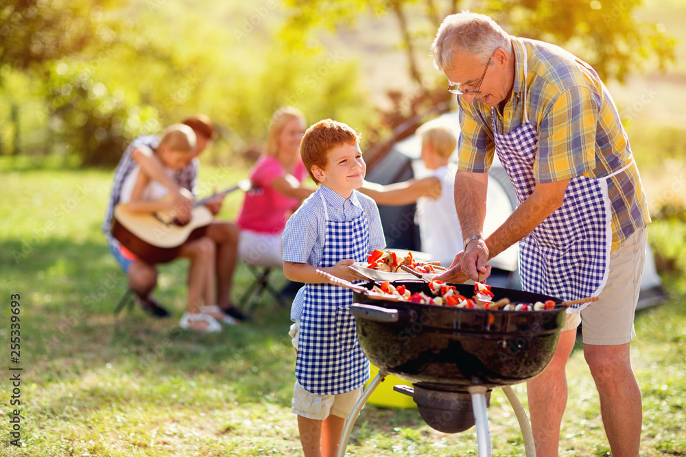 smiling grandfather giving grandson grilling meat. - obrazy, fototapety, plakaty 