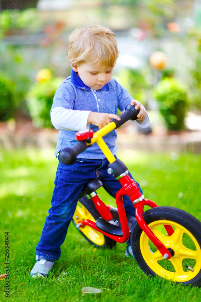 Active blond kid boy in colorful clothes driving balance and learner's bike or bicycle in domestic garden. Toddler child dreaming and having fun on warm summer day. outdoors movement game for children Active blond kid boy in colorful clothes driving balance and learner's bike or bicycle in domestic garden. Toddler child dreaming and having fun on warm summer day. outdoors movement game for children