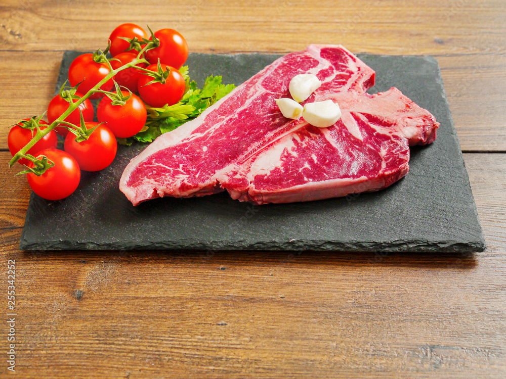 Raw T-bone steak on a stone plate with baby tomato on a vine and garlic cloves, wooden table. Raw T-bone steak on a stone plate with baby tomato on a vine and garlic cloves, wooden table.