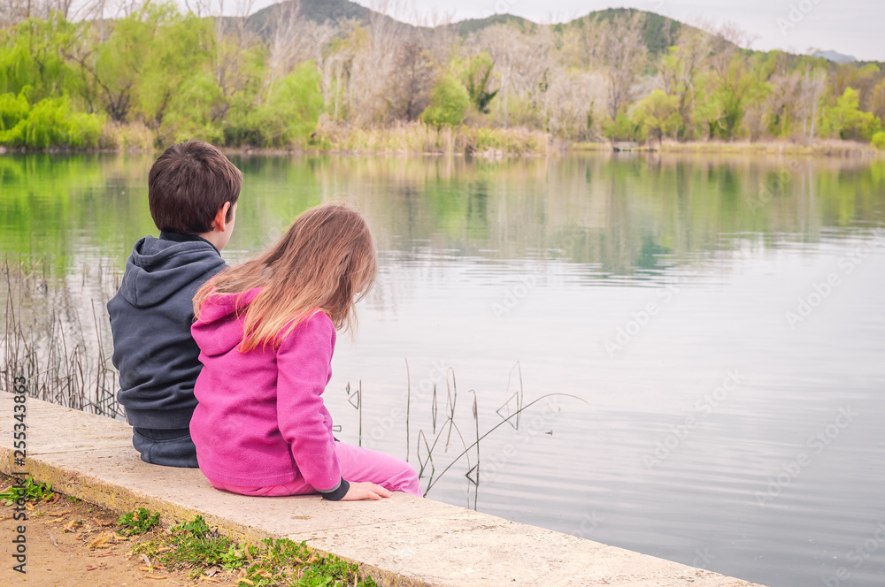 Children sit by the lake and admire in spring nature. Unity with nature. Children relax outdoors. Freedom and unity concept. Children sit by the lake and admire in spring nature. Unity with nature. Children relax outdoors. Freedom and unity concept.