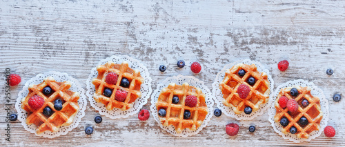 Traditional waffles with fresh raspberries and blueberries on lace doily on wooden background. Top view, copy space, flat lay .