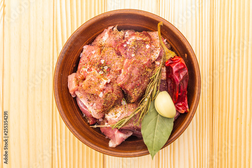 Raw pork shoulder. Spices, dry herbs, garlic, rosemary, bay leaf, hot pepper, pepper mix, Ingredients for marinating meat. Rustic ceramic bowl on wooden background, top view, close up.