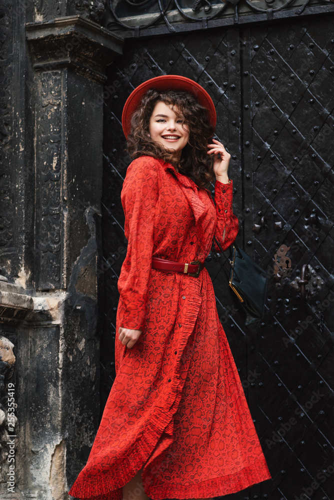 Outdoor portrait of young beautiful happy smiling fashionable lady wearing orange hat, snakeskin printed midi dress, posing in street