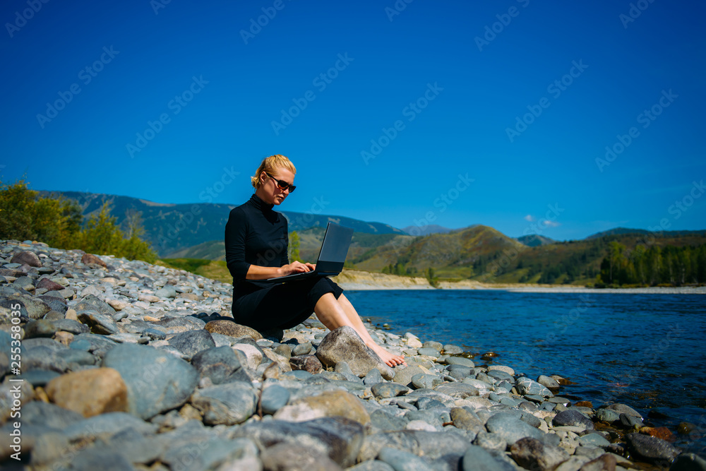 Freelancer with laptop on the nature. Blogger hipster traveler business lady pretty girl  works outdoor. Young blondy woman in black sitting next to mountain river