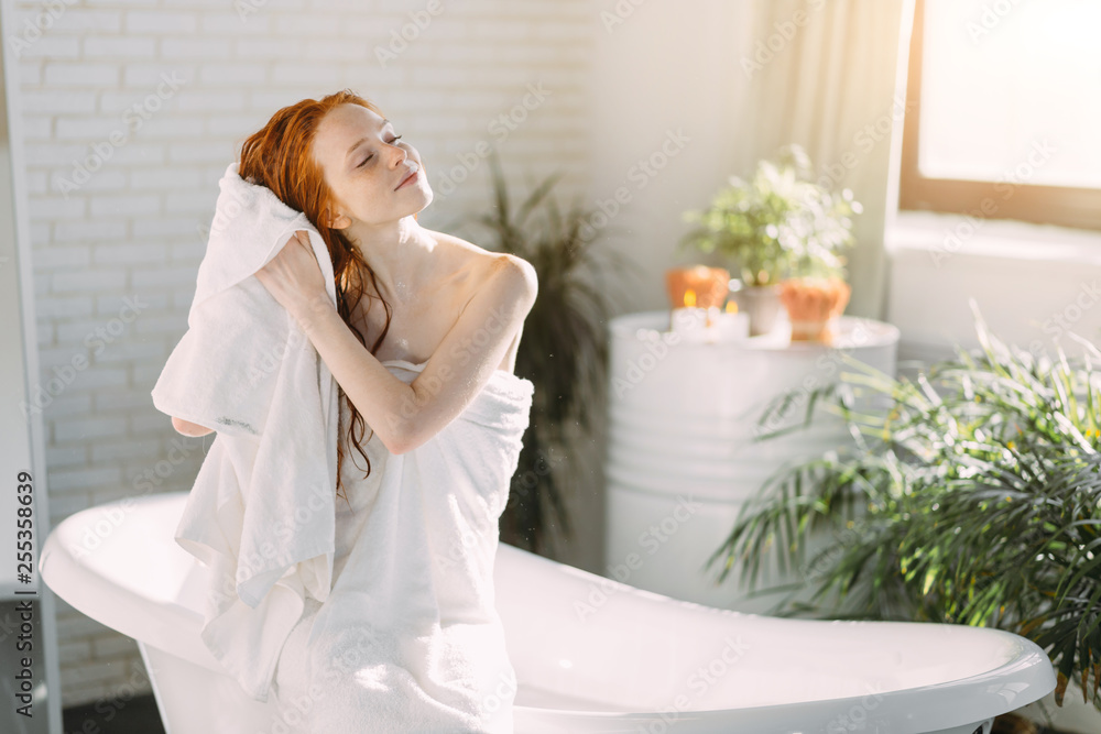 Restful relaxing redhead caucasian young woman drying her coppery hair with a towel after a healing balm, sitting on the rim of a bathtub in spa centre. Restful relaxing redhead caucasian young woman drying her coppery hair with a towel after a healing balm, sitting on the rim of a bathtub in spa centre.