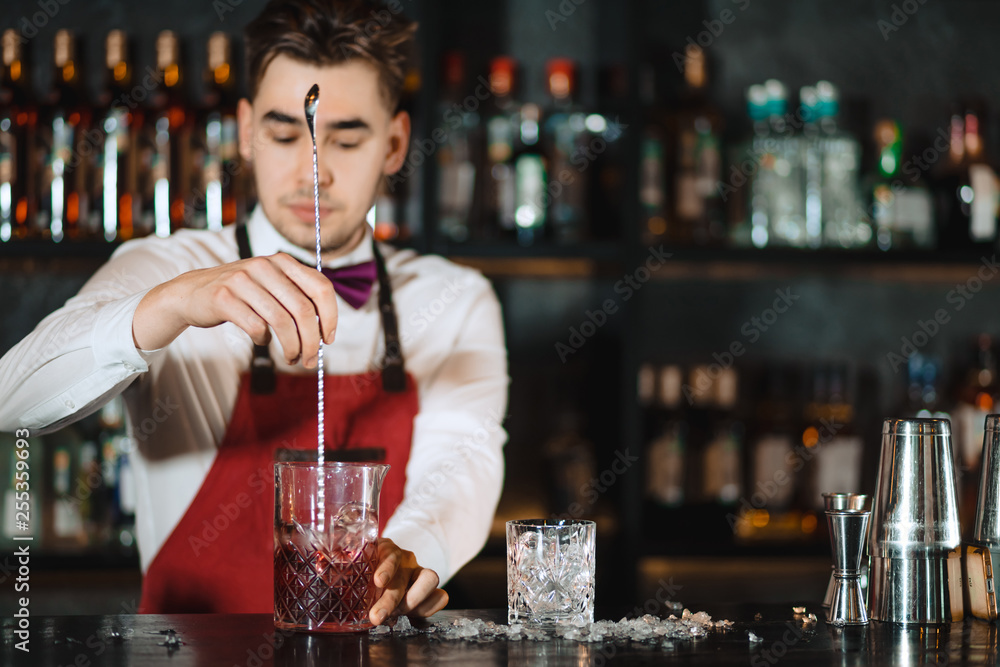 Young professional bartender mixing with long metal spoon cocktail in a glass standing at wooden bar counter with bottles of alcohol, blurry background.