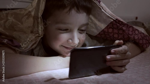 A boy lying on a bed at home, relaxing, smiling and watching video on smartphone in the dark under the blanket