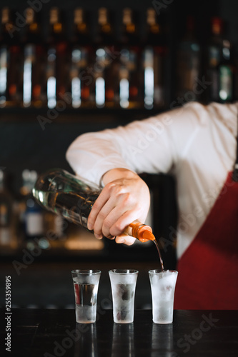 Expert barman in elegant uniform working at night party at popular nightclub, pouring gin or vodka in frosted short glasses