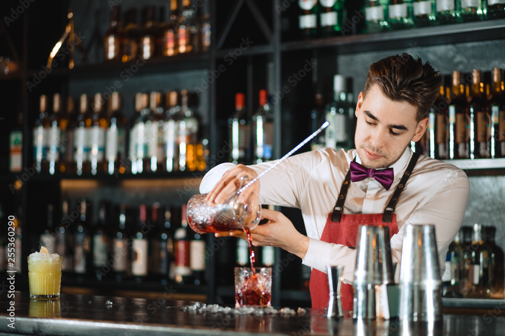 Professional bartender in uniform doing show of his work, holding two parts of metal shaker in his hands and pouring a cocktail, shelves full of bottles with alcohol on the background