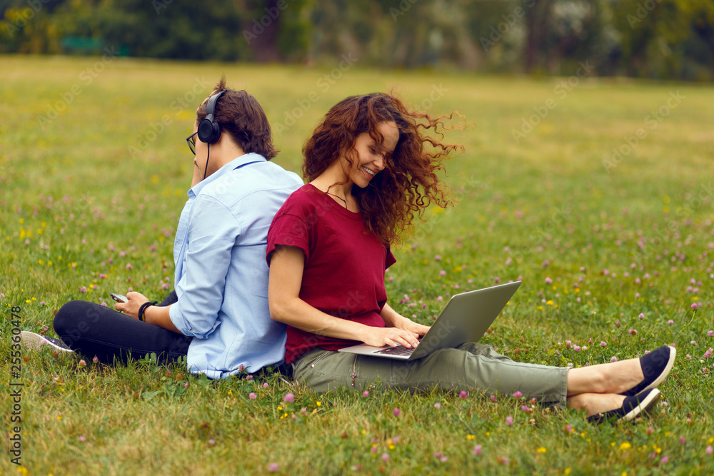 Image of a two young person, sitting behind, using laptop and listening music in headphone, isolated on a green grass in open air. Leisure time together. Horizontal view. Image of a two young person, sitting behind, using laptop and listening music in headphone, isolated on a green grass in open air. Leisure time together. Horizontal view.