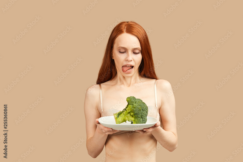 Skinny woman with ginger long hair, dressed only in underwear, looking at piece of fresh broccoli with adoration, licking, wants to eat it very much Skinny woman with ginger long hair, dressed only in underwear, looking at piece of fresh broccoli with adoration, licking, wants to eat it very much