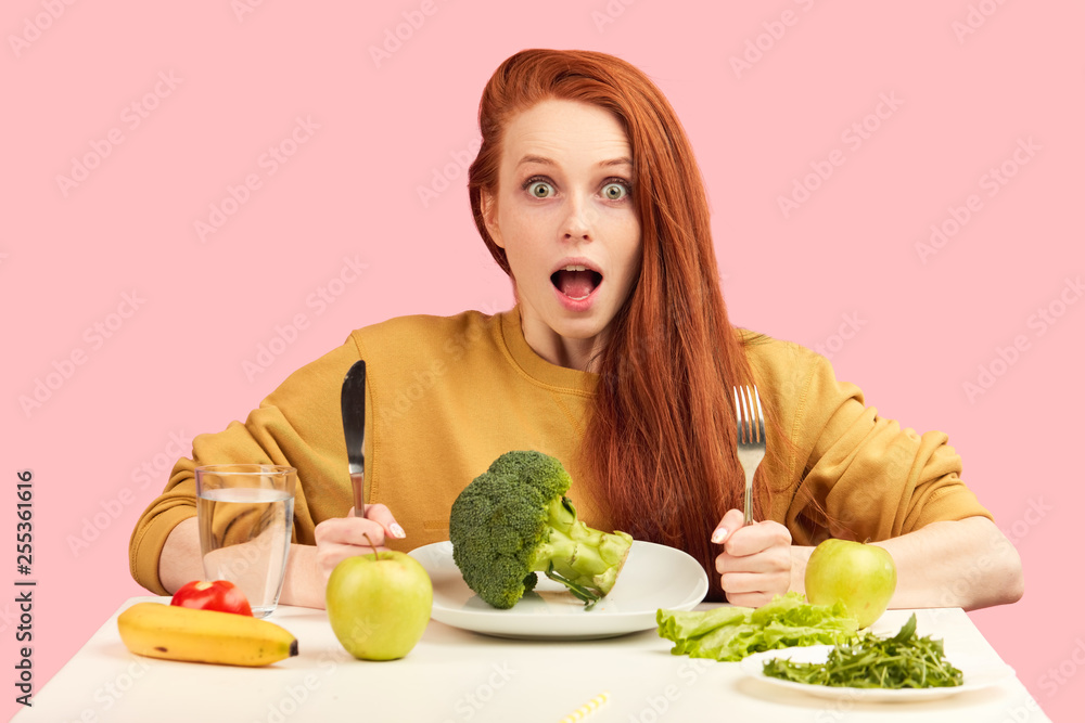 Healthy eating and dieting and calories concept. Surprised woman with wide opened eyes and mouth looking at camera with astonishment, sitting at table with fresh vegetables