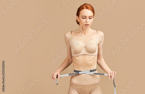 Young red-haired caucasian skinny woman is measuring her waist during diet, standing undressed over beige studio background. Diet Concept. Weight Control