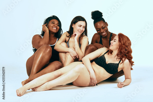 Studio portrait of four multicultural young women of different ethnicity and size, smiling cheerfully, talking with each other, sitting over white background