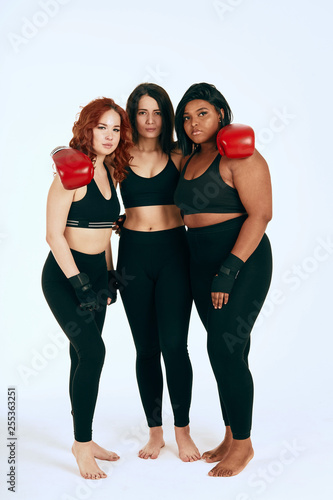 Group of three multiracial diverse woman in black sportswear posing in boxing gloves after workout over white background, smiling cheerfully at camera