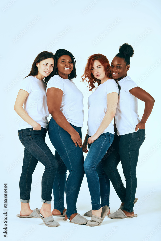 Four young multi ethnic female models of different race, body size and hair color smiling at camera, posing on white studio background in white casual t-shirts and skinny jeans. Four young multi ethnic female models of different race, body size and hair color smiling at camera, posing on white studio background in white casual t-shirts and skinny jeans.