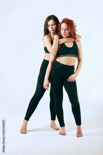 Homosexual couple of caucasian young brunette and red haired women standing side by side in black sportive clothes, tank top and leggings in studio over white background