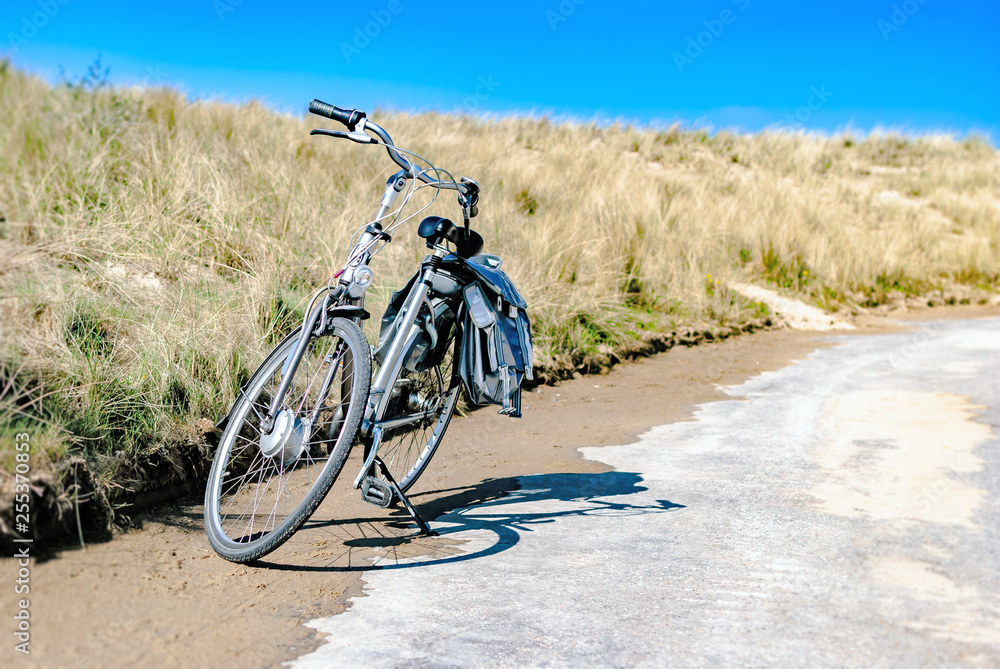 Fototapeta premium A bicycle parked along a coastal road with dunes at the background. Summer concept. Vacation. Bike rental. Biking. Tourism. Retreat. Spring. Nature therapy. Vlissingen, the Netherlands. Northern sea.