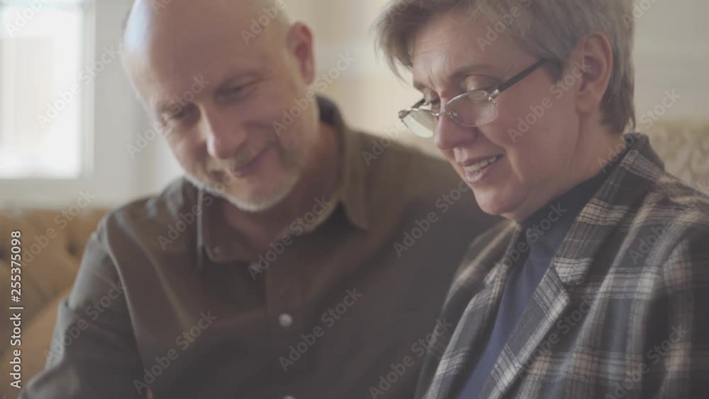 Portrait of mature couple, man and woman, sitting on the sofa with a book and talking and smiling to each other. Family relationships. S-log, ungraded Portrait of mature couple, man and woman, sitting on the sofa with a book and talking and smiling to each other. Family relationships. S-log, ungraded