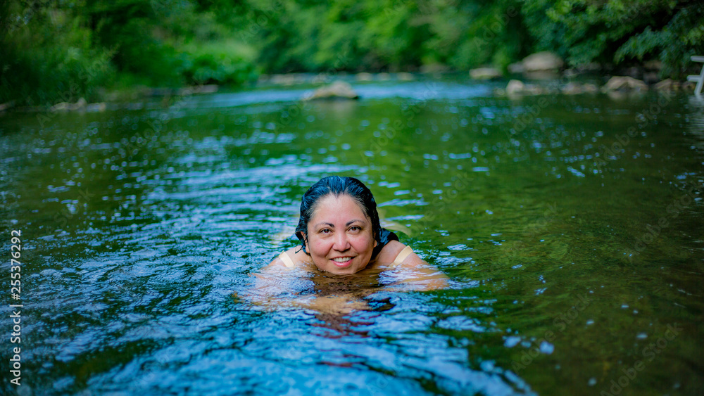 River with a Mexican woman swimming with green vegetation in a blurred background, looking at the camera, long wet wavy hair and no makeup. Wonderful sunny summer day in Schuld, Germany