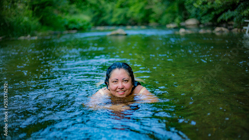 River with a Mexican woman swimming with green vegetation in a blurred background, looking at the camera, long wet wavy hair and no makeup. Wonderful sunny summer day in Schuld, Germany