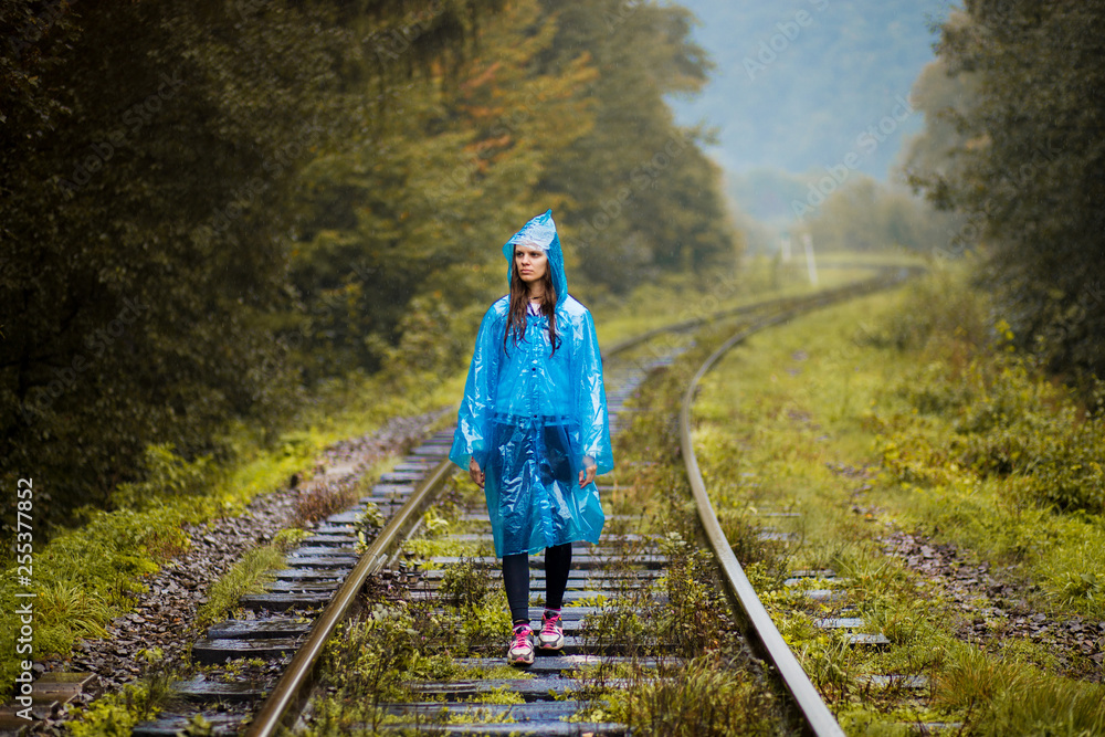 Girl traveller wearing blue jacket and go by forest railway. Autumn and raining season with dark green tones while girl in blue rain jacket walks in forest, fog, rain and clouds