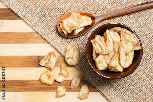 Banana chips inside brown ceramic and wooden spoon on bamboo wooden background.