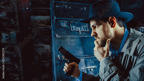 Detective with the gun wearing a fedora hat and a trench coat, dark background.