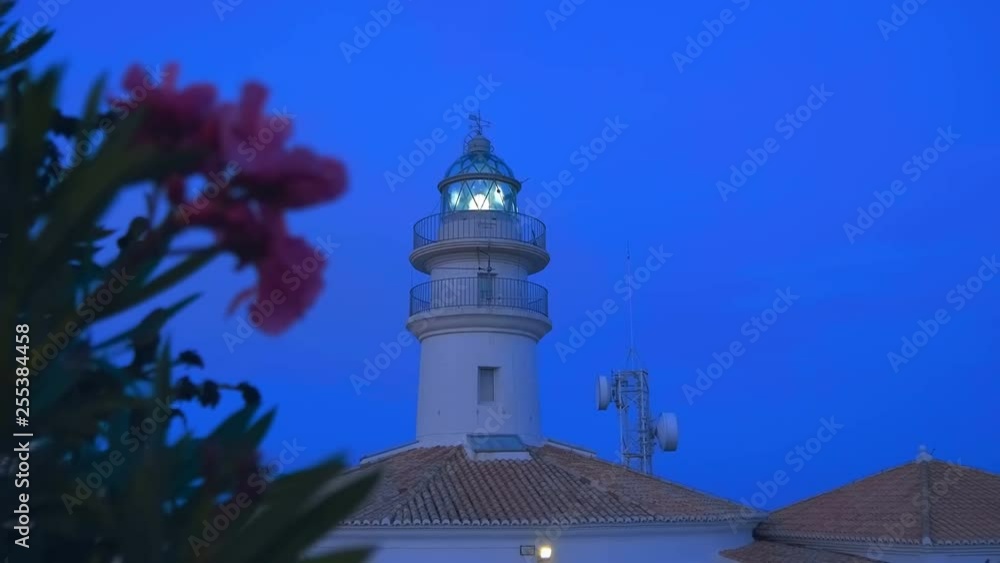 The lighthouse is shining by sector lights and telecommunication tower with satellite dishes stands on a cliff on the shores of the Mediterranean Sea against the night blue sky The lighthouse is shining by sector lights and telecommunication tower with satellite dishes stands on a cliff on the shores of the Mediterranean Sea against the night blue sky