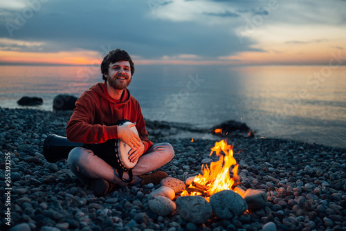 percussionist guy plays a djembe sitting on beach by the fire at sunset.