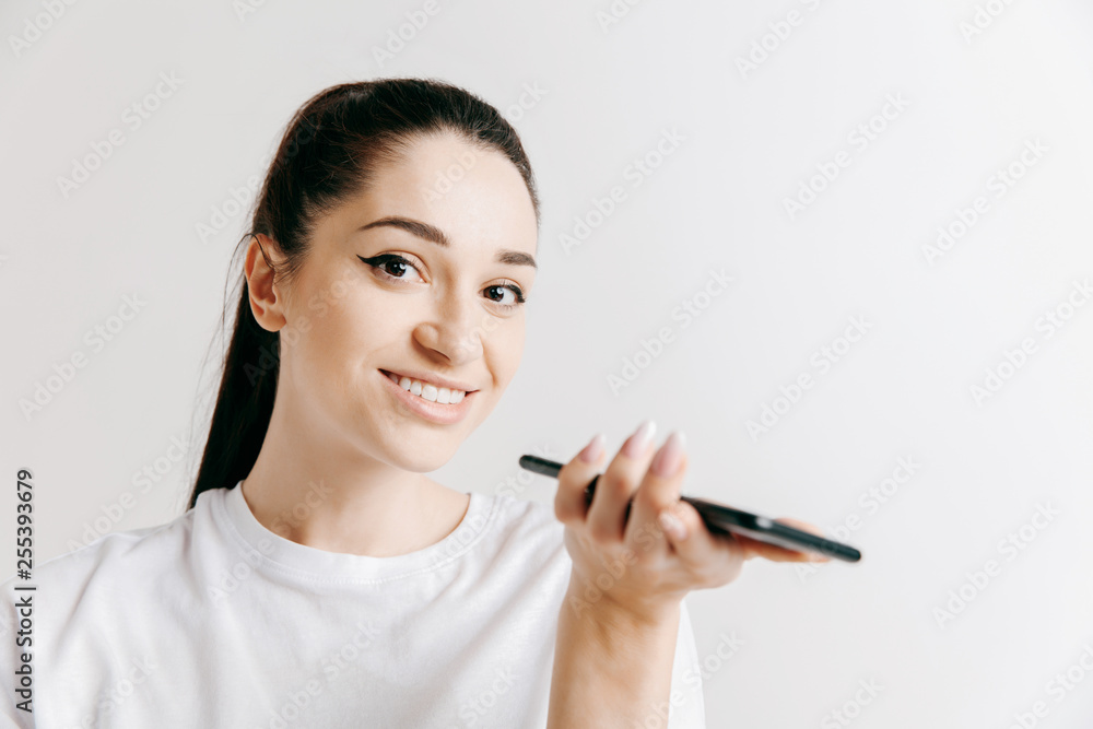 Indoor portrait of attractive young woman isolated on gray background, holding blank smartphone, using voice control, feeling happy and surprised. Human emotions, facial expression concept. Indoor portrait of attractive young woman isolated on gray background, holding blank smartphone, using voice control, feeling happy and surprised. Human emotions, facial expression concept.
