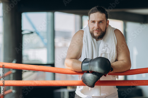 Sweaty sportive boxer wearing boxing bandages on hands looking on camera from ring while having break after workout in gym
