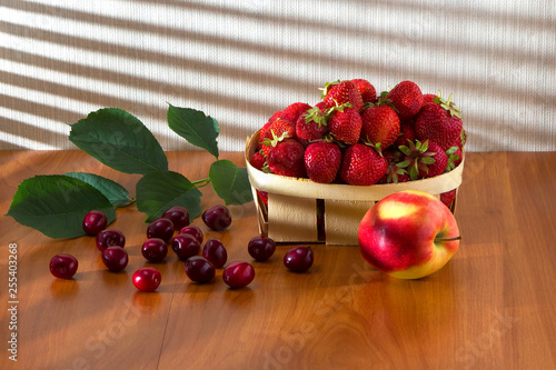 Juicy, ripe cherries lie on a table with green foliage under the rays of a bright sun, close by, an apple and strawberries in a basket.